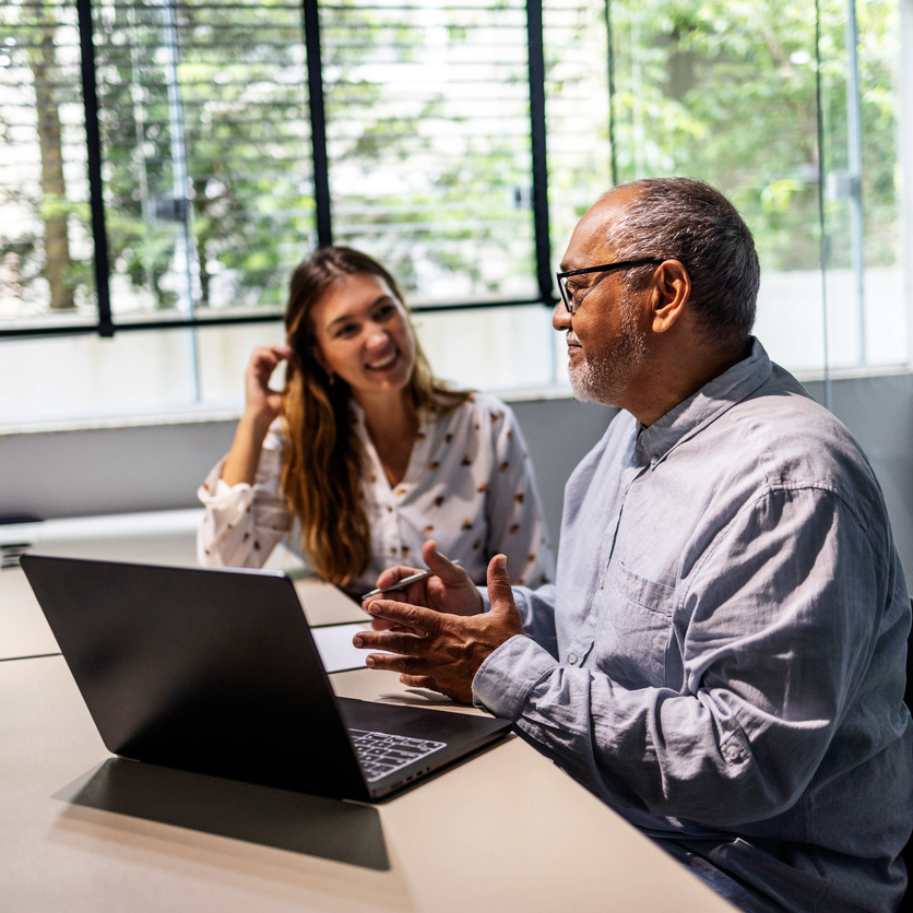 A younger woman and an older man sitting in front of laptop talking and smiling